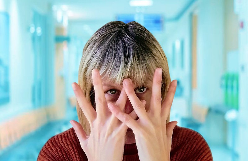 Woman with dark hair and red jumper with hands stretched over face, in front of blue blurry hospital corridor.