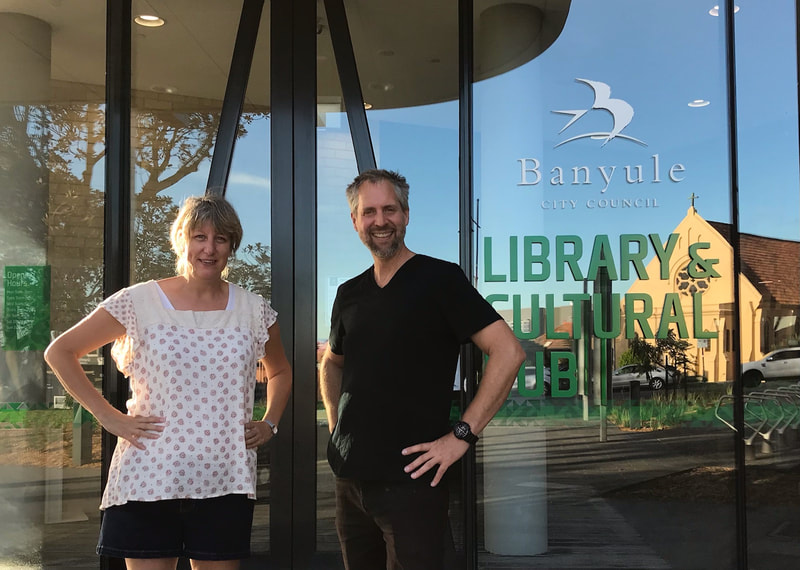 Smiling woman in white top with smiling man in black top, standing in front of glass wall with Banyule Library and Cultural Hub on it, sunny day.