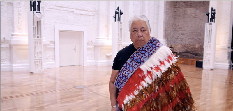 Woman of Maori descent wearing a korowai, a garment woven with red and white feathers. She is standing inside a big white hall with hardwood floor.